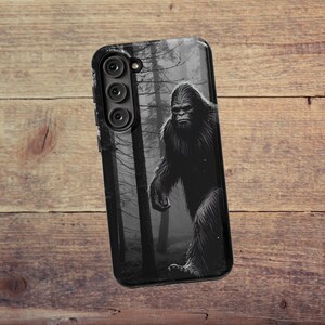 May include: Black phone case with a graphic of Bigfoot walking through a dark forest. The phone case has a triple camera lens.