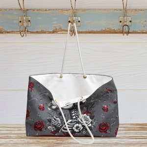 May include: A large gray tote bag with a white skeleton and floral design. The bag has red roses and white flowers with a gray background. The bag has white rope handles.
