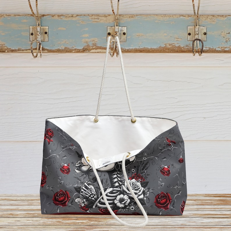 May include: A large gray tote bag with a white skeleton and floral design. The bag has red roses and white flowers with a gray background. The bag has white rope handles.