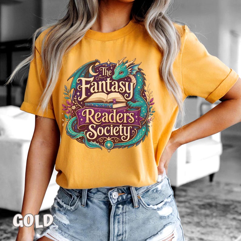 May include: A gold-colored short-sleeve t-shirt featuring a graphic print of a teal dragon curled around an open book. The text reads The Fantasy Readers Society in a decorative font, surrounded by floral elements and a crescent moon.