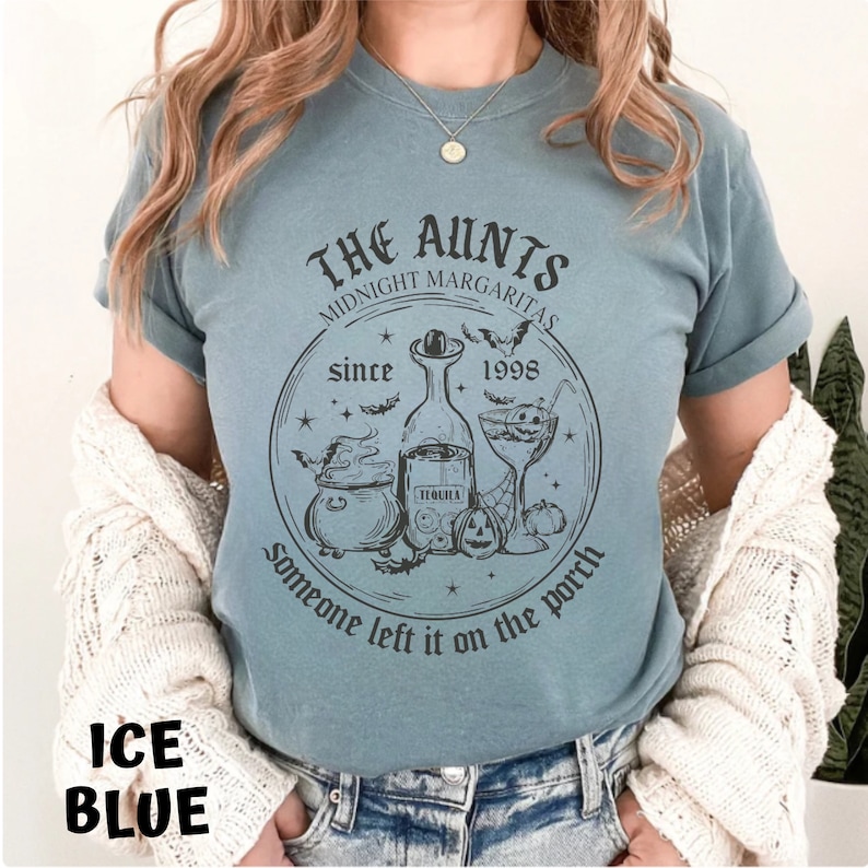 K&ouml;nnte beinhalten: Ein hellblaues T-Shirt mit einem schwarzen Grafikdesign, das einen Kessel, eine Flasche Tequila, ein Cocktailglas und Flederm&auml;use zeigt. Der Text "THE AUNTS MIDNIGHT MARGARITAS since 1998 someone left it on the porch" ist auf das T-Shirt gedruckt.