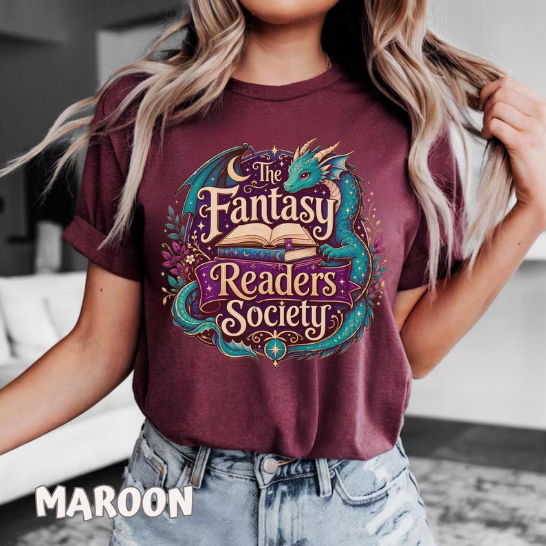May include: A maroon t-shirt featuring a graphic print of a teal dragon coiled around an open book. The text reads The Fantasy Readers Society in a decorative font. The design includes stars, crescent moons, and floral elements in purple and gold.