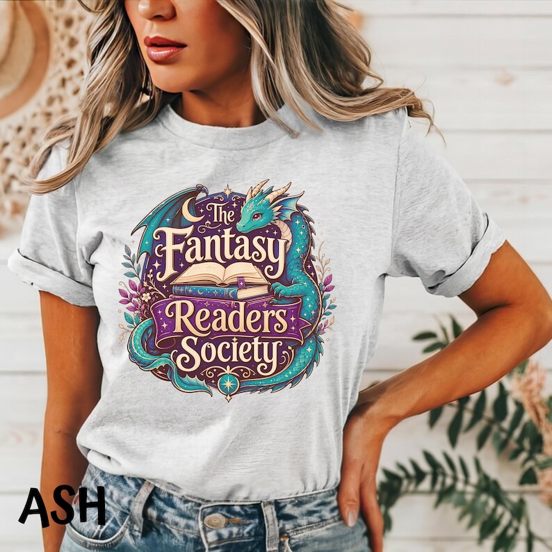 May include: A light gray short-sleeve t-shirt featuring a graphic print of a teal dragon curled around an open book. The text reads The Fantasy Readers Society in stylized purple and cream lettering, surrounded by stars and floral elements.