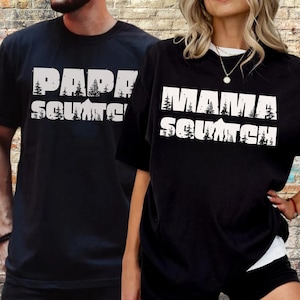 May include: Two black t-shirts with white text. The shirt on the left says "Papa Squatch" and the shirt on the right says "Mama Squatch".