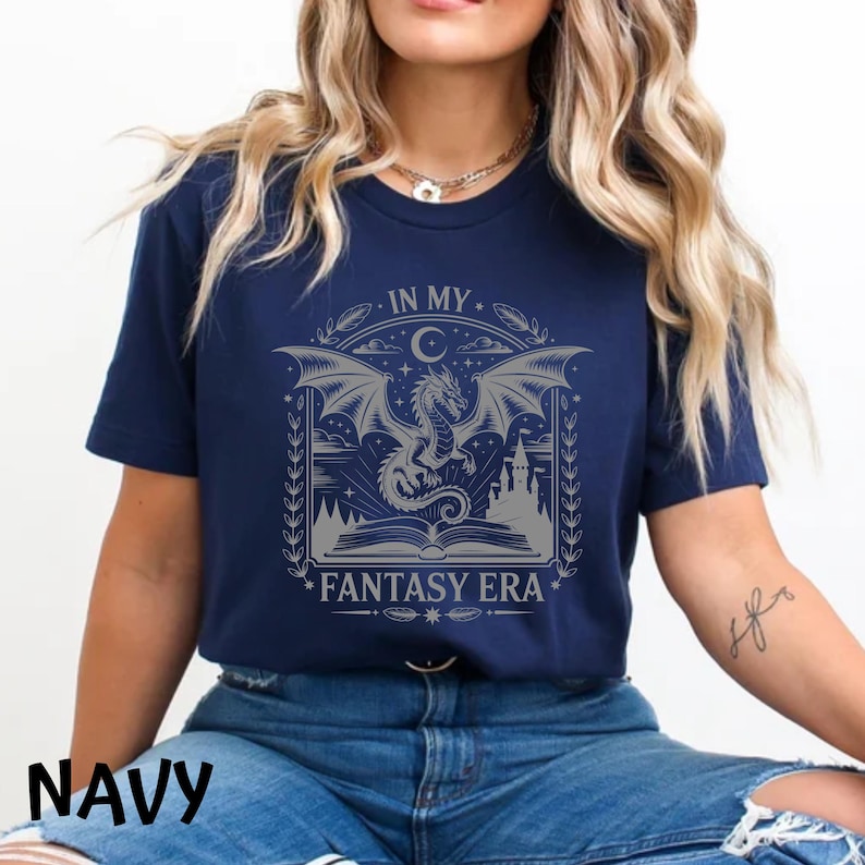 May include: A navy blue short-sleeve t-shirt featuring a light gray graphic print of a dragon rising from an open book, with a castle silhouette, stars, and a crescent moon. The text reads IN MY FANTASY ERA in a decorative, stylized font.