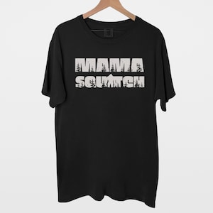 May include: Black t-shirt with a white graphic print that says "Mama Squatch" with a mountain and trees in the background.