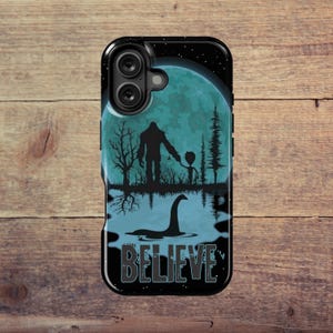 May include: Black phone case featuring a Bigfoot silhouette holding hands with an alien, with a lake scene and a Loch Ness Monster. A full moon and the word "BELIEVE" are also featured in the design.