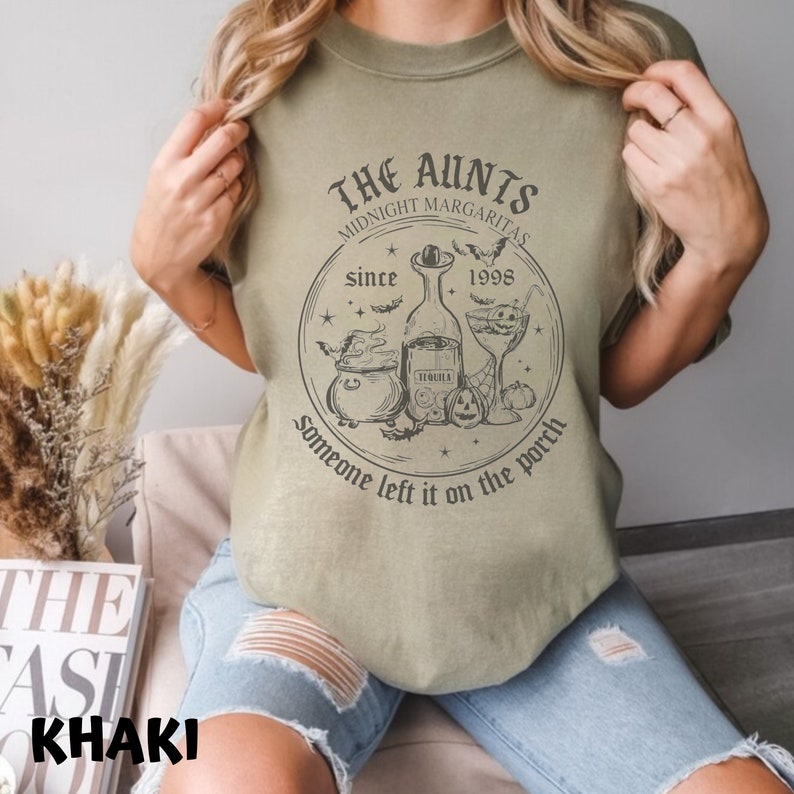 K&ouml;nnte beinhalten: Ein khakifarbenes T-Shirt mit einem schwarzen grafischen Design, das einen Kessel, eine Flasche Tequila, ein Margarita-Glas und Flederm&auml;use zeigt. Der Text auf dem Shirt lautet "The Aunts Midnight Margaritas since 1998 someone left it on the porch".