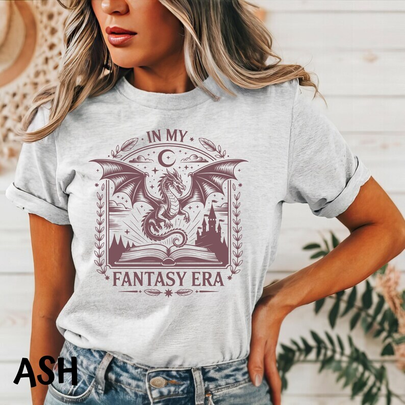 May include: A light gray crew neck t-shirt featuring a dark maroon graphic print of a dragon flying above an open book and a castle silhouette. The text "IN MY FANTASY ERA" is printed in a decorative font above and below the central illustration.