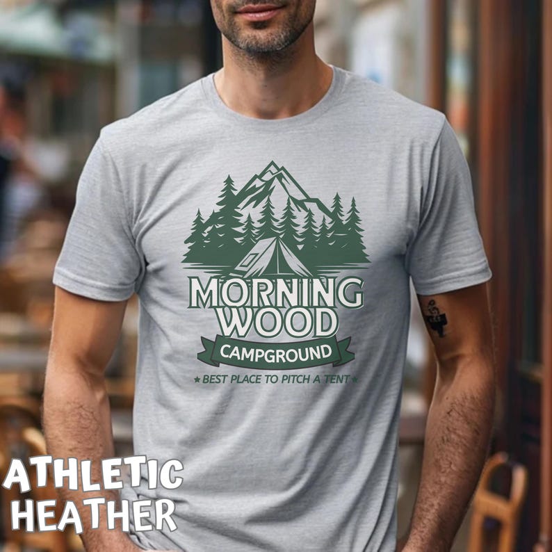 Puede incluir: Una camiseta gris jaspeada con un gr&aacute;fico verde de una cordillera, &aacute;rboles y una tienda de campa&ntilde;a. El texto "Morning Wood Campground" est&aacute; impreso en la camiseta, con el lema "Best place to pitch a tent".