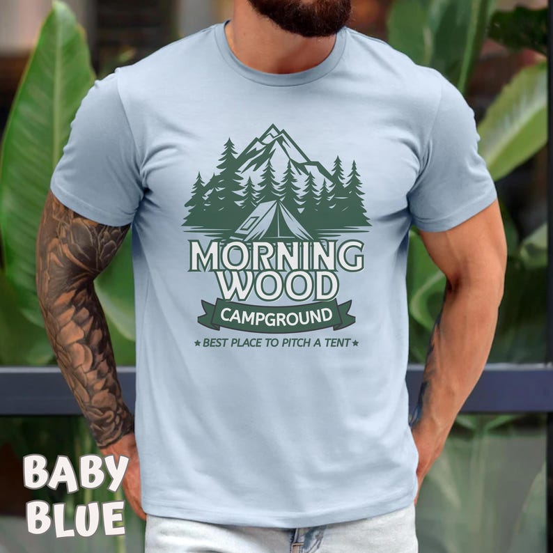 Puede incluir: Una camiseta azul claro con un gr&aacute;fico verde de una cordillera, &aacute;rboles y una tienda de campa&ntilde;a. El texto "Morning Wood Campground" est&aacute; impreso en la camiseta, junto con el lema "Best place to pitch a tent."