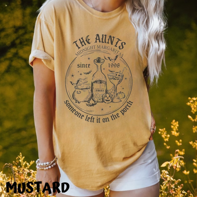 K&ouml;nnte beinhalten: Ein senfgelbes T-Shirt mit einem schwarzen grafischen Design, auf dem "The Aunties Midnight Margaritas since 1998 Someone left it on the porch" steht. Die Grafik zeigt einen Kessel, eine Flasche Tequila, ein Margarita-Glas und Flederm&auml;use.