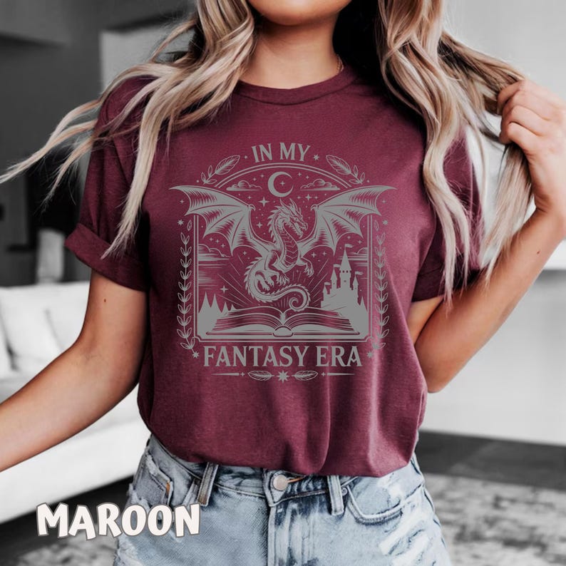 May include: A maroon short-sleeve t-shirt featuring a light gray graphic print of a dragon emerging from an open book with a castle in the background, a crescent moon, and the text "IN MY FANTASY ERA" centered within a decorative frame.