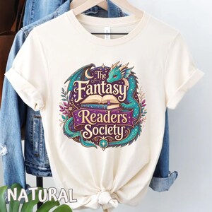 May include: A natural-colored t-shirt featuring a graphic of a teal dragon curled around an open book. The text reads The Fantasy Readers Society in purple and cream lettering, surrounded by decorative leaves, stars, and a crescent moon.