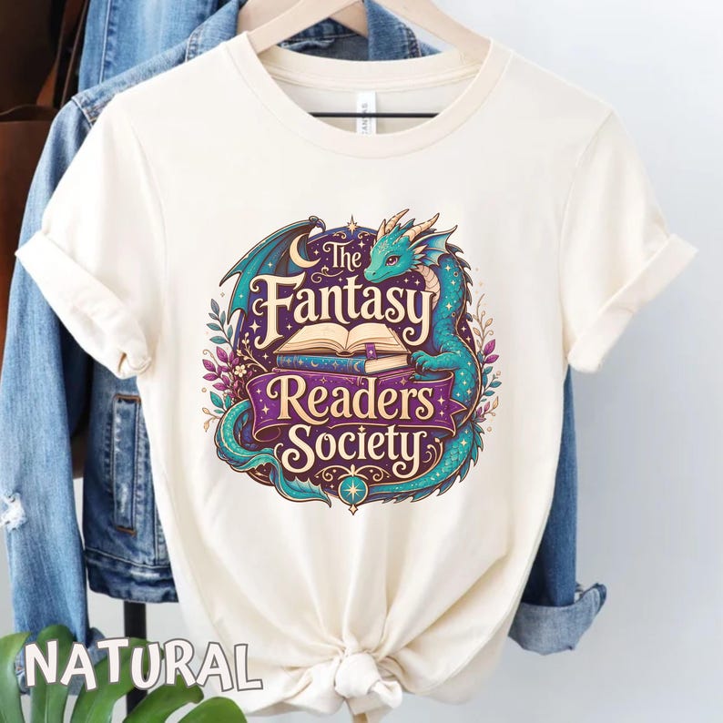 May include: A natural-colored t-shirt featuring a graphic of a teal dragon curled around an open book. The text reads The Fantasy Readers Society in purple and cream lettering, surrounded by decorative leaves, stars, and a crescent moon.