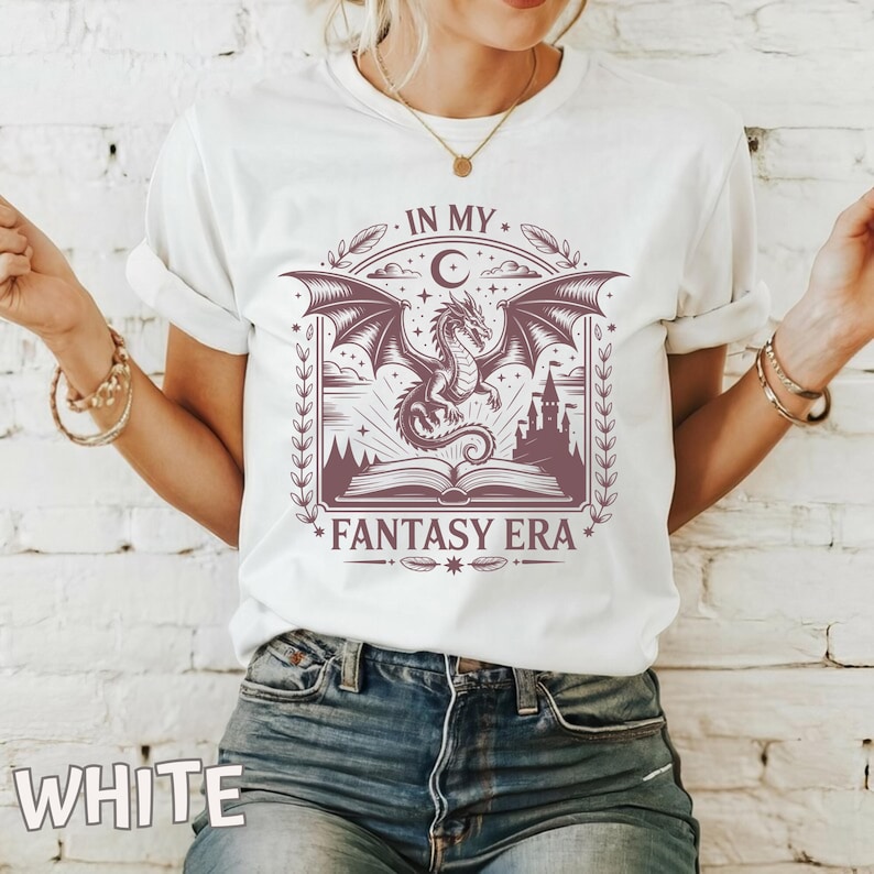May include: A white short-sleeve t-shirt featuring a dark purple graphic print of a dragon emerging from an open book with a castle in the background. The design includes the text "IN MY FANTASY ERA" framed by decorative leaves, stars, and a crescent moon.