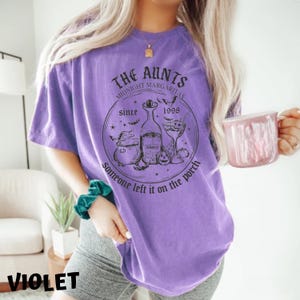 K&ouml;nnte beinhalten: Lilafarbenes T-Shirt mit einem schwarzen grafischen Design, auf dem "The Aunts Midnight Margaritas Since 1998 Someone left it on the porch" steht. Die Grafik zeigt einen Kessel, eine Flasche Tequila, ein Glas Margarita und eine Fledermaus.