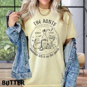 K&ouml;nnte beinhalten: Ein gelbes T-Shirt mit einem schwarzen grafischen Design, auf dem "The Aunts Midnight Margaritas Since 1998 Someone left it on the porch" steht. Die Grafik enth&auml;lt einen Kessel, eine Flasche Tequila, ein Cocktailglas und Flederm&auml;use.