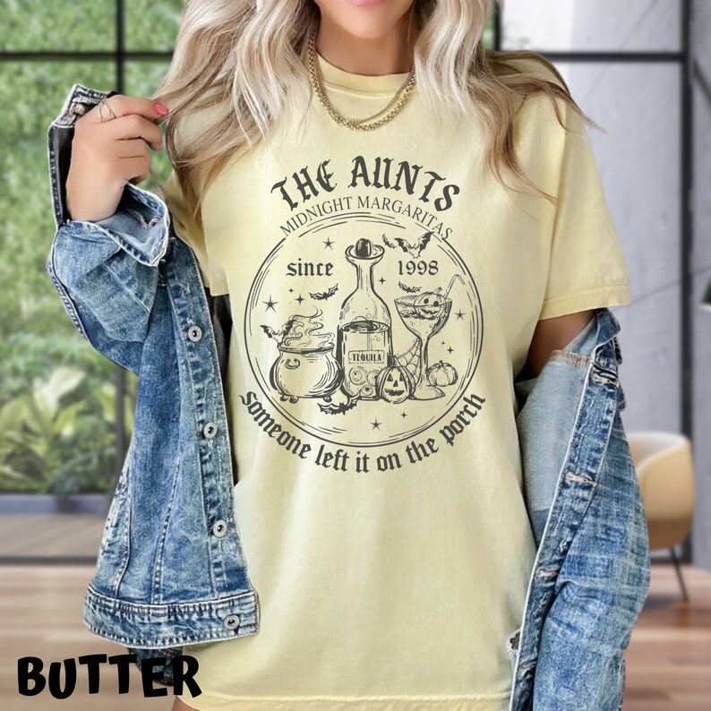 K&ouml;nnte beinhalten: Ein gelbes T-Shirt mit einem schwarzen grafischen Design, auf dem "The Aunts Midnight Margaritas Since 1998 Someone left it on the porch" steht. Die Grafik enth&auml;lt einen Kessel, eine Flasche Tequila, ein Cocktailglas und Flederm&auml;use.