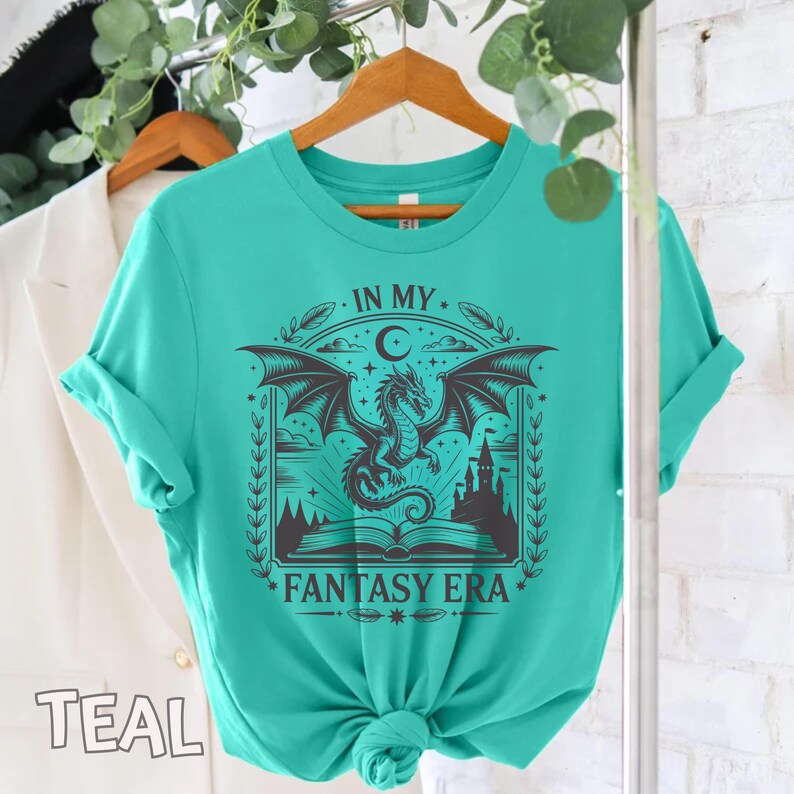 May include: A teal short-sleeve t-shirt featuring a black graphic print of a dragon flying above an open book and a castle. The text "IN MY FANTASY ERA" is printed in black above and below the illustration, which is framed by leaves.