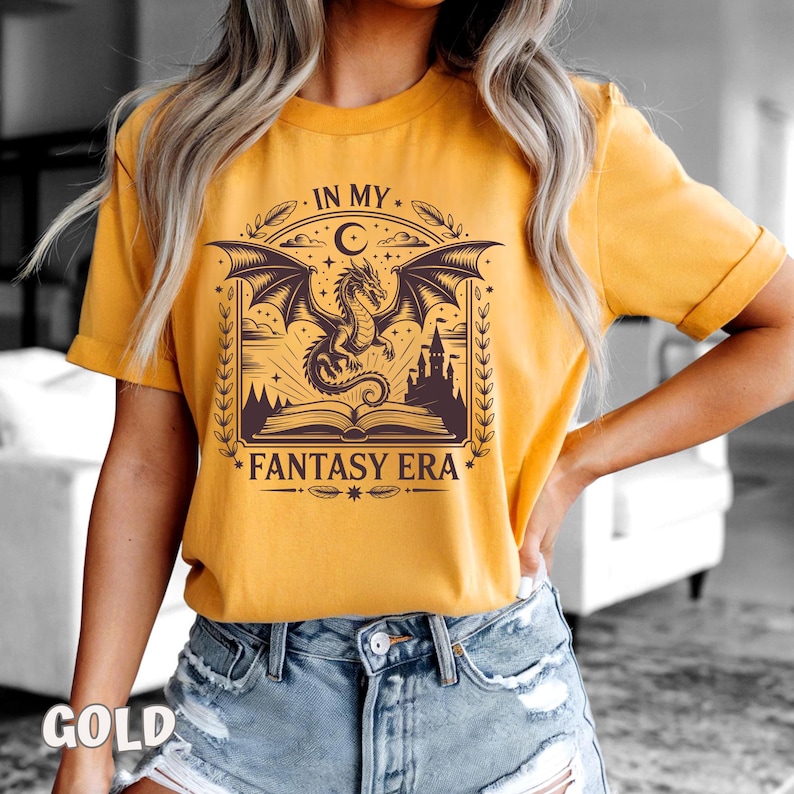 May include: A mustard yellow short-sleeve t-shirt featuring a dark brown graphic print of a dragon flying above an open book and a castle. The text "IN MY FANTASY ERA" is printed in a decorative font above and below the central illustration.