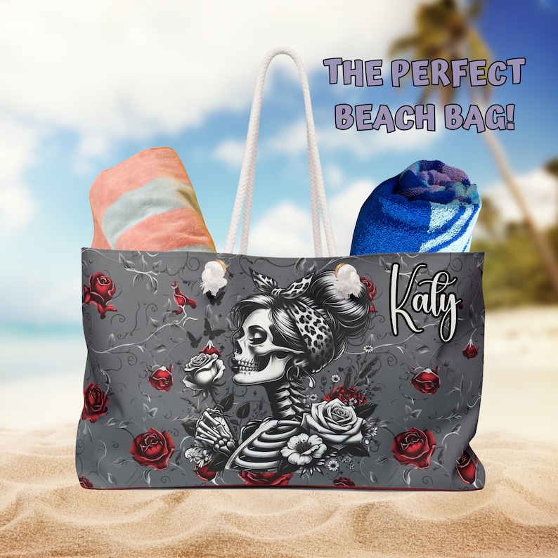 May include: A gray beach tote bag with a floral design featuring a skeleton woman with a rose in her hand. The bag has a white rope handle and the name "Katy" is printed on the side.