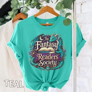 May include: A teal t-shirt featuring a graphic print of a dragon curled around an open book. The text reads The Fantasy Readers Society in a decorative font. The design includes purple banners, small stars, and floral elements in shades of blue.