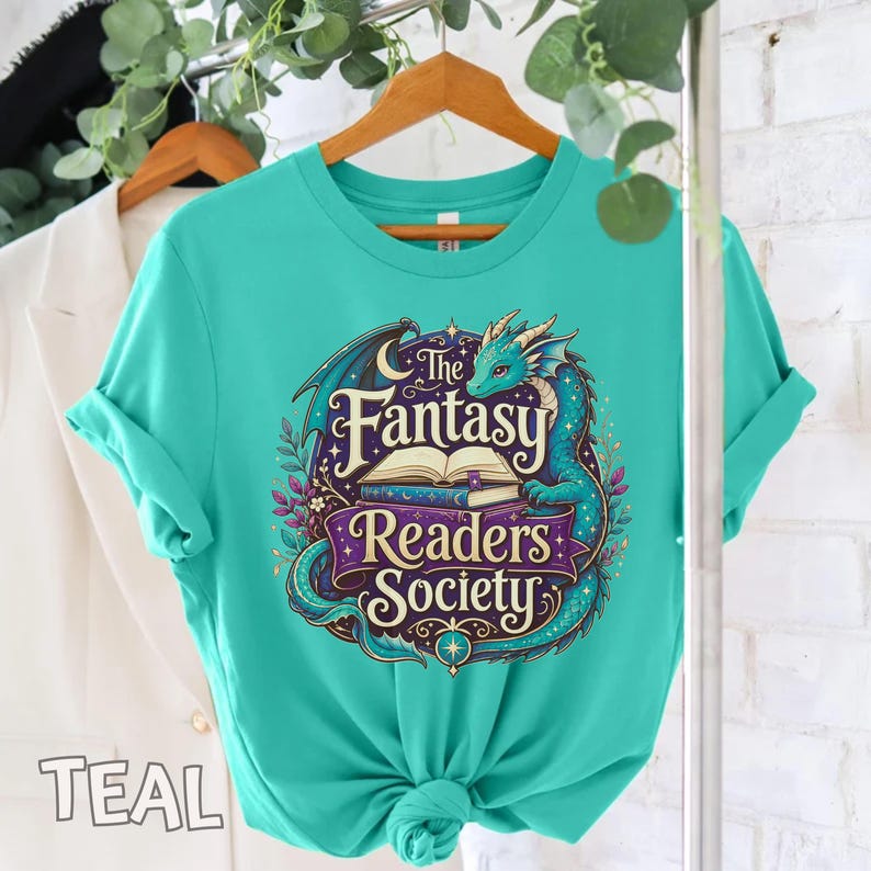 May include: A teal t-shirt featuring a graphic print of a dragon curled around an open book. The text reads The Fantasy Readers Society in a decorative font. The design includes purple banners, small stars, and floral elements in shades of blue.
