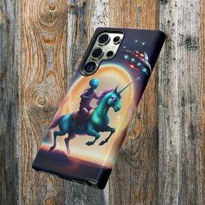 May include: A phone case with a colorful illustration of an alien riding a unicorn in front of a moon and a flying saucer. The phone case is black and has a camera lens cutout.