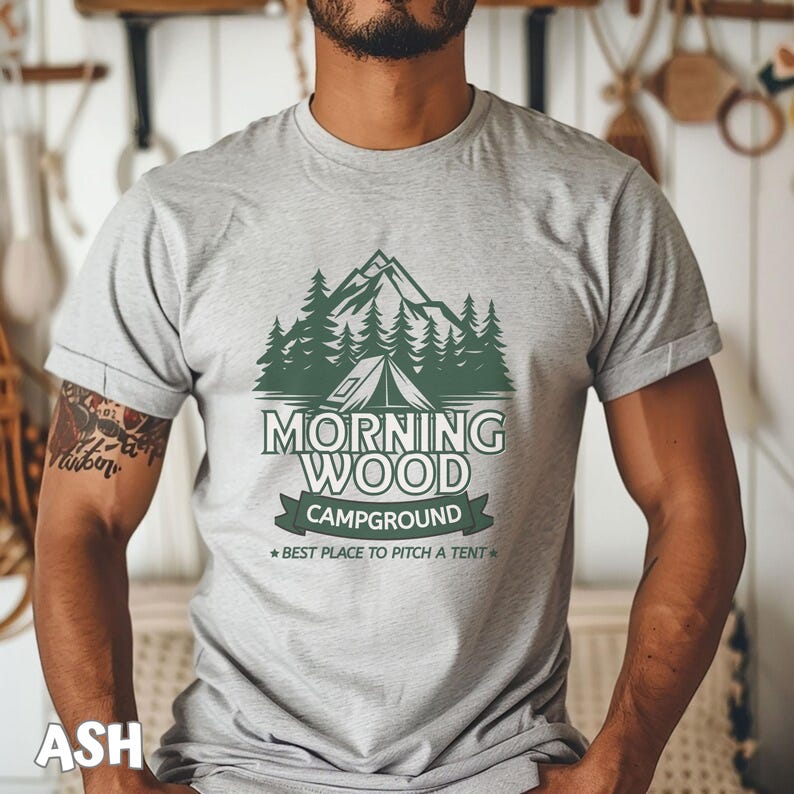 Puede incluir: Una camiseta gris con un gr&aacute;fico verde de una cordillera, &aacute;rboles y una tienda de campa&ntilde;a. El texto "Morning Wood Campground" est&aacute; impreso en la camiseta, junto con la frase "Best place to pitch a tent."