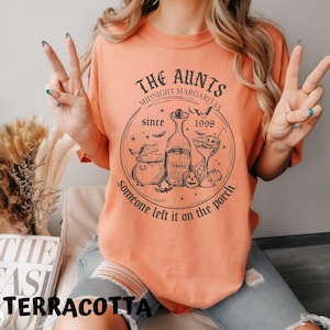 K&ouml;nnte beinhalten: Ein terracottafarbenes T-Shirt mit einem schwarzen Grafikdesign. Die Grafik zeigt einen Kessel, eine Flasche Tequila, ein Cocktailglas, Flederm&auml;use und den Text "The Aunts Midnight Margaritas since 1998 someone left it on the porch".