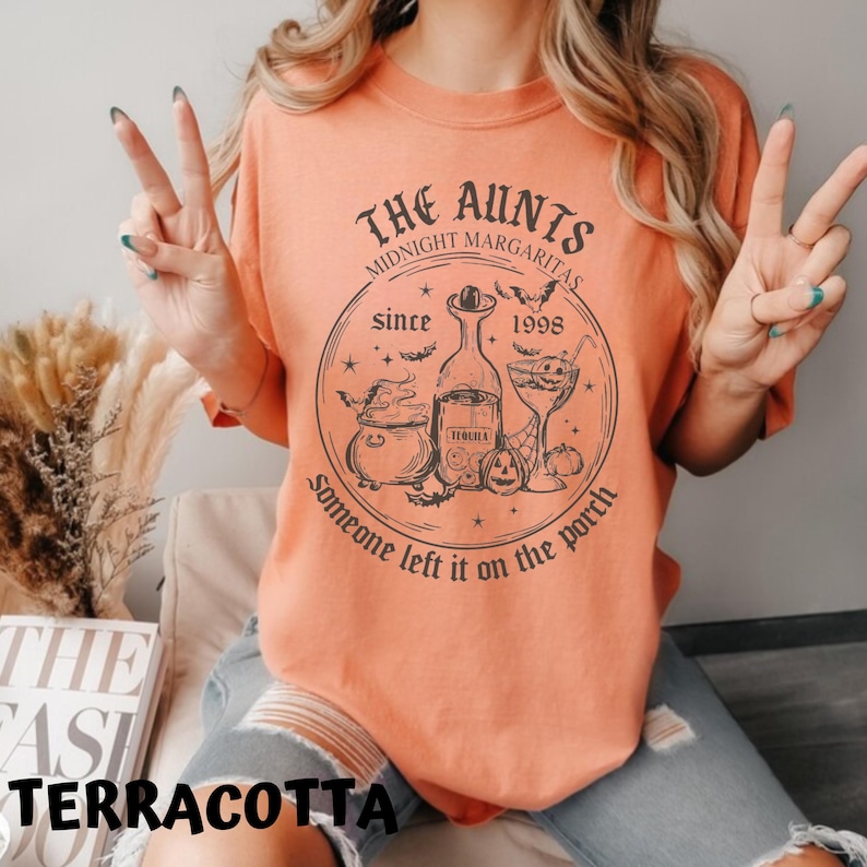 K&ouml;nnte beinhalten: Ein terracottafarbenes T-Shirt mit einem schwarzen Grafikdesign. Die Grafik zeigt einen Kessel, eine Flasche Tequila, ein Cocktailglas, Flederm&auml;use und den Text "The Aunts Midnight Margaritas since 1998 someone left it on the porch".