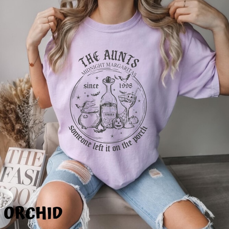 K&ouml;nnte beinhalten: Ein lila T-Shirt mit einem schwarzen grafischen Design, auf dem "The Aunts Midnight Margaritas since 1998 someone left it on the porch" steht. Die Grafik zeigt einen Kessel, eine Flasche Tequila, ein Cocktailglas und Flederm&auml;use.