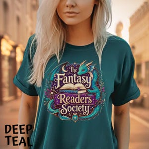May include: A deep teal t-shirt featuring a graphic print of a turquoise dragon curled around an open book. The text reads The Fantasy Readers Society in a decorative font, surrounded by stars, crescent moons, and floral elements in purple and gold.