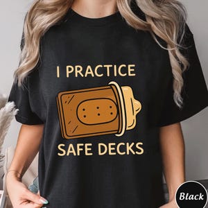 May include: Black t-shirt with the text "I PRACTICE SAFE DECKS" above a graphic of a skateboard deck with a bandage. The graphic is in shades of brown and yellow.