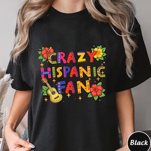 May include: Black t-shirt featuring the text "CRAZY HISPANIC FAN" in vibrant colours. The design incorporates a guitar and floral elements in red, yellow, and green. A casual wear item.