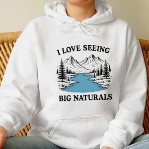 May include: White hoodie with a graphic of a mountain landscape, a river, and pine trees. The text "I LOVE SEEING BIG NATURALS" is printed in black above and below the image.