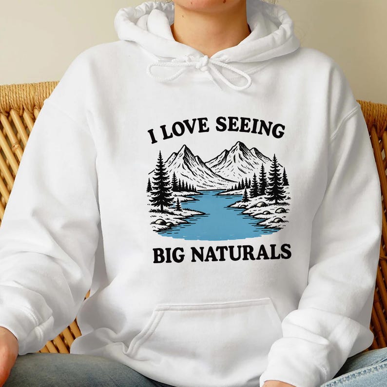 May include: White hoodie with a graphic of a mountain landscape, a river, and pine trees. The text "I LOVE SEEING BIG NATURALS" is printed in black above and below the image.