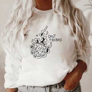 Pu&ograve; includere: Felpa bianca con grafica nera di un dado a venti facce avvolto dalle fiamme. Il testo "CAST FIREBALL" &egrave; scritto sopra il dado. Un top casual a maniche lunghe.