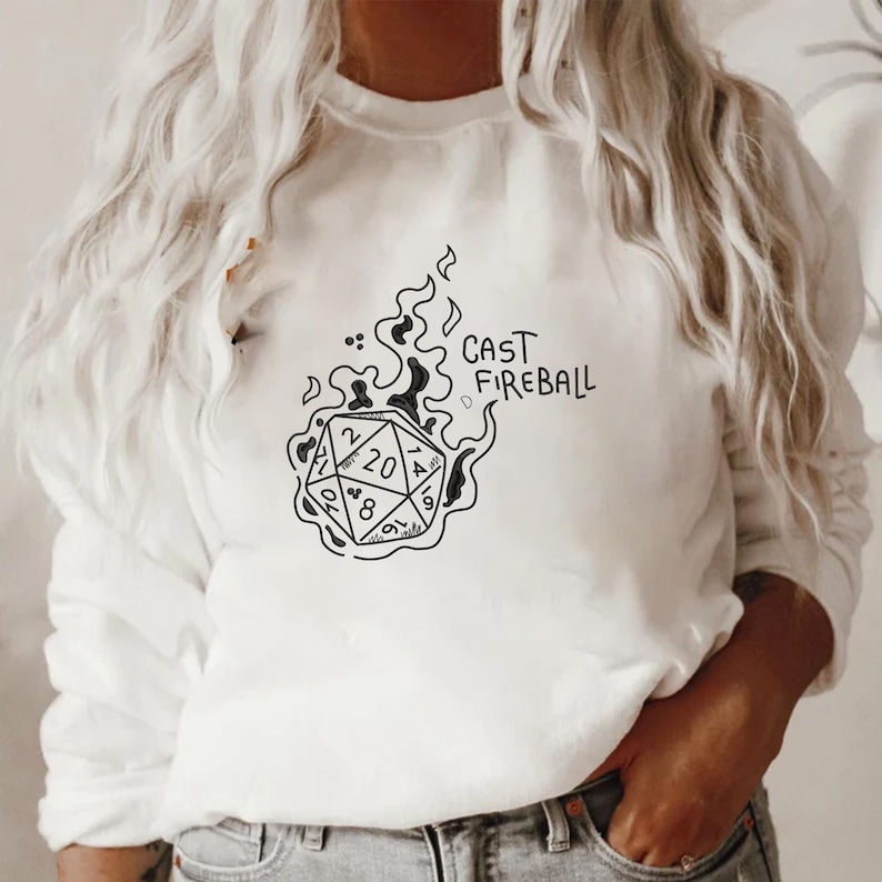 Pu&ograve; includere: Felpa bianca con grafica nera di un dado a venti facce avvolto dalle fiamme. Il testo "CAST FIREBALL" &egrave; scritto sopra il dado. Un top casual a maniche lunghe.