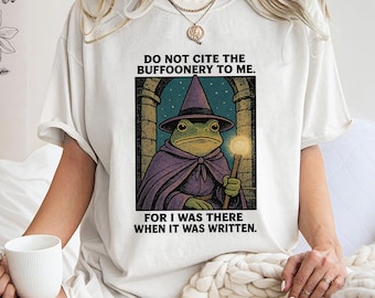Op de afbeelding: Crèmekleurig T-shirt met een illustratie van een kikker-tovenaar in een paarse cape en hoed, met een gloeiende staf. De tekst "DO NOT CITE THE BUFFOONERY TO ME. FOR I WAS THERE WHEN IT WAS WRITTEN" staat boven en onder de afbeelding.