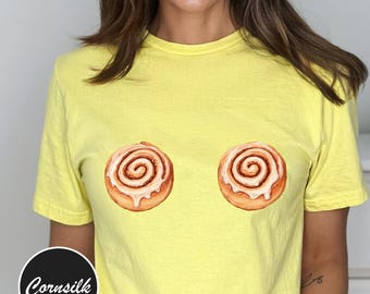 May include: A yellow t-shirt featuring two watercolor-style cinnamon roll graphics. The shirt has short sleeves and a round neckline. The brand name "Cornsilk" is printed on a black and white circular logo on the bottom left.