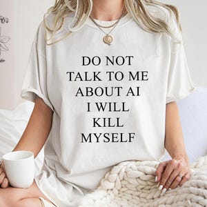 May include: Cream-colored t-shirt with the text "DO NOT TALK TO ME ABOUT AI I WILL KILL MYSELF" in black. The shirt has short sleeves and a relaxed fit. The person is wearing a gold necklace and holding a white mug.