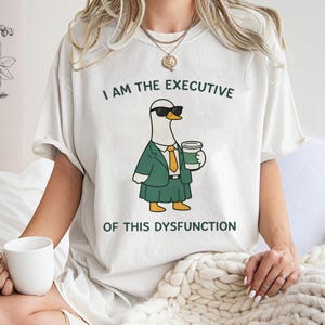 Może przedstawiać: Kremowa koszulka z kreskówkową gęsią w okularach przeciwsłonecznych, zielonym garniturze i kubku z kawą. Tekst brzmi "I AM THE EXECUTIVE OF THIS DYSFUNCTION" w kolorze zielonym.
