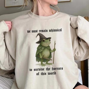 Peut inclure: Sweat-shirt beige avec une illustration fantaisiste d'une grenouille sorcier tenant un bâton et une épée. Le texte "we must remain whimsical to survive the horrors of this world" est imprimé au-dessus et en dessous de la grenouille.