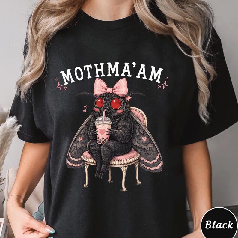 May include: A black t-shirt featuring a graphic of a fuzzy black moth with large red eyes, wearing a pink bow, sitting on a small chair, and holding a bubble tea cup. The text MOTHMA'AM is printed in white above the moth, with small pink hearts and stars.