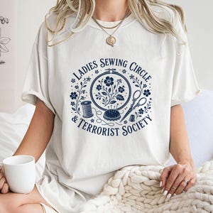 May include: Cream-colored t-shirt with a navy blue graphic. The graphic features a circular design with the text "LADIES SEWING CIRCLE & TERRORIST SOCIETY" surrounding a floral and sewing-themed illustration. Includes a spool of thread, scissors, and a pin cushion.