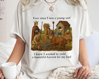 Può includere: T-shirt color crema con un'illustrazione di una scena agricola medievale e testo. La grafica raffigura persone che lavorano in un campo con una mucca, una casa e un re. Il testo recita: "Da quando ero un giovane servo, sapevo che volevo produrre un raccolto abbondante per il mio signore."