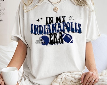 Könnte beinhalten: Weißes T-Shirt mit einer blauen Grafik, auf der "In My Indianapolis Era" mit einem Fußball und einem Helm steht.
