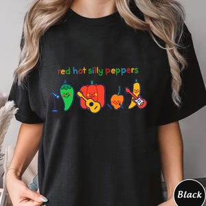 Pu&ograve; includere: Maglietta nera con il testo "red hot silly peppers" in colori arcobaleno. Il testo &egrave; sopra una linea di cinque peperoni dei cartoni animati che suonano strumenti musicali.